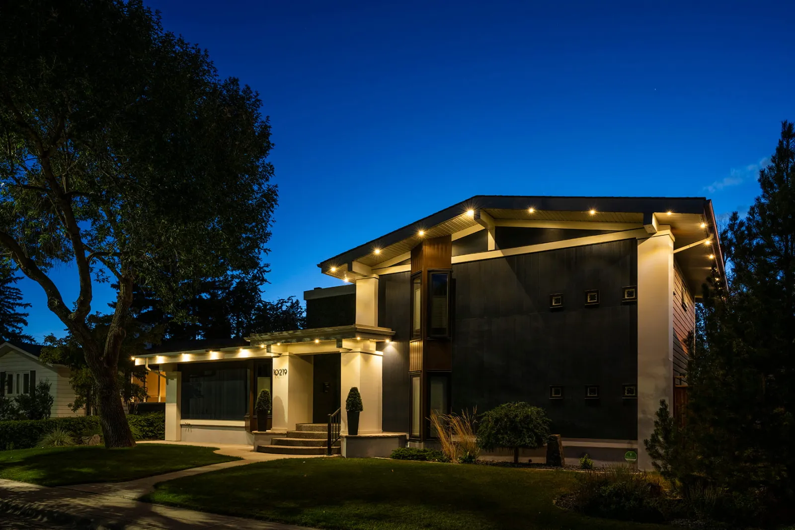 Modern two-story home with warm downlights along its flat roof edges at dusk