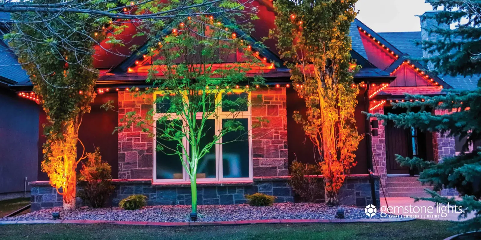 Brick home at night with trees in the front yard lit orange and green by landscape lights