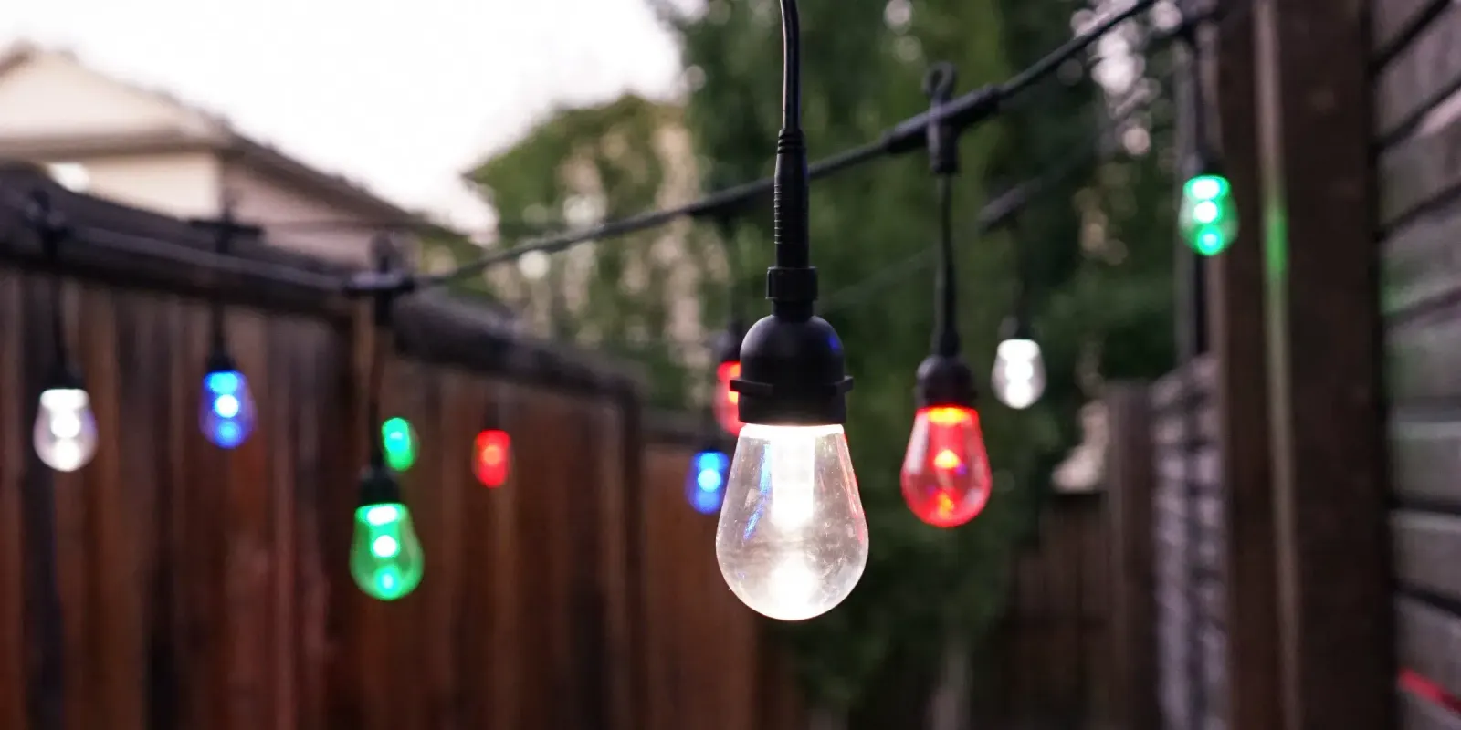 Patio string lights with colored and white bulbs running along a backyard fence at dusk