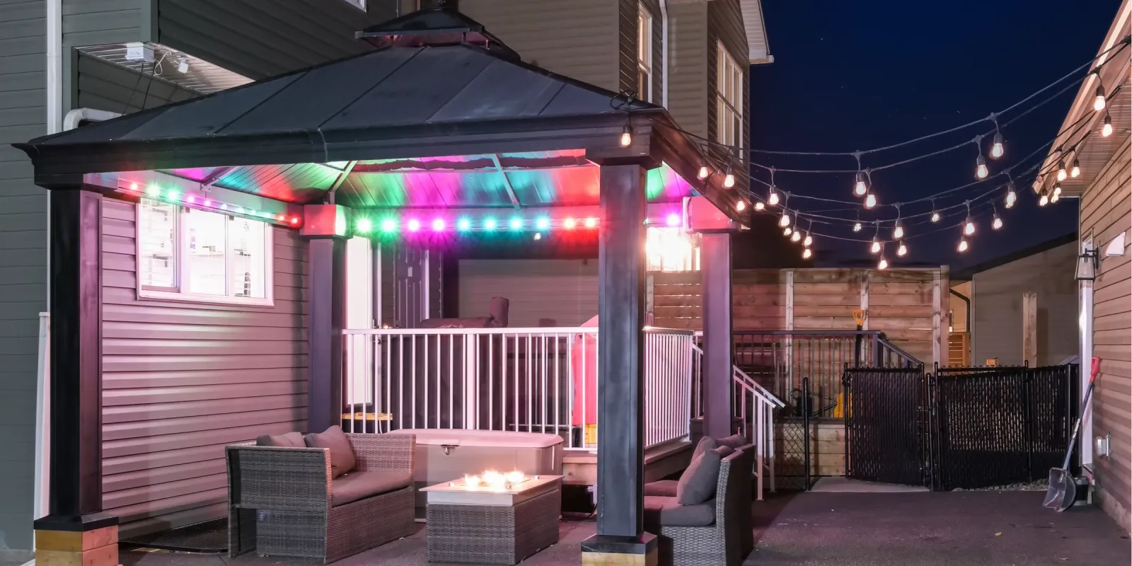 Backyard gazebo trimmed with colored smart lights beside a run of patio string lights