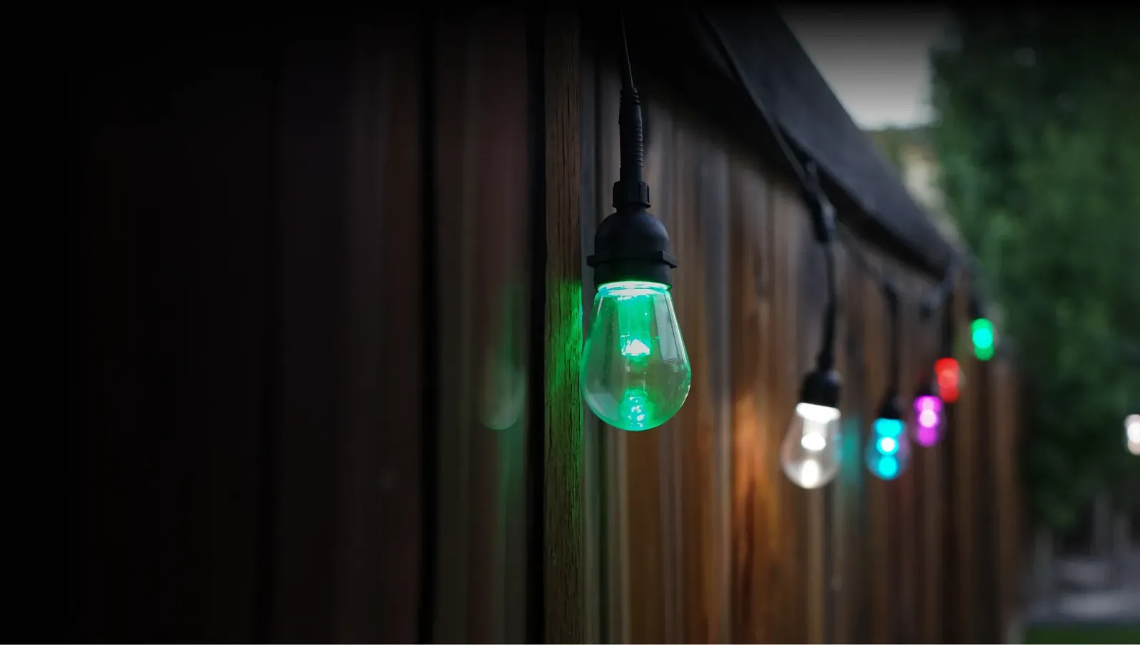 Smart string light bulbs glowing green, white, blue and red along a wooden fence at dusk