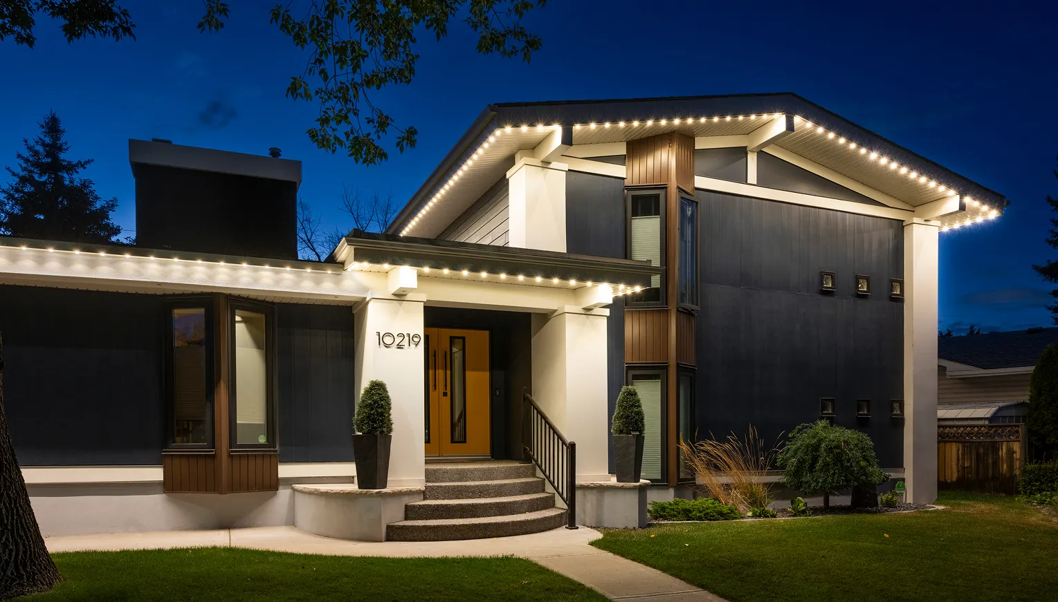 Modern two-story home at dusk with warm white lights tracing every roofline