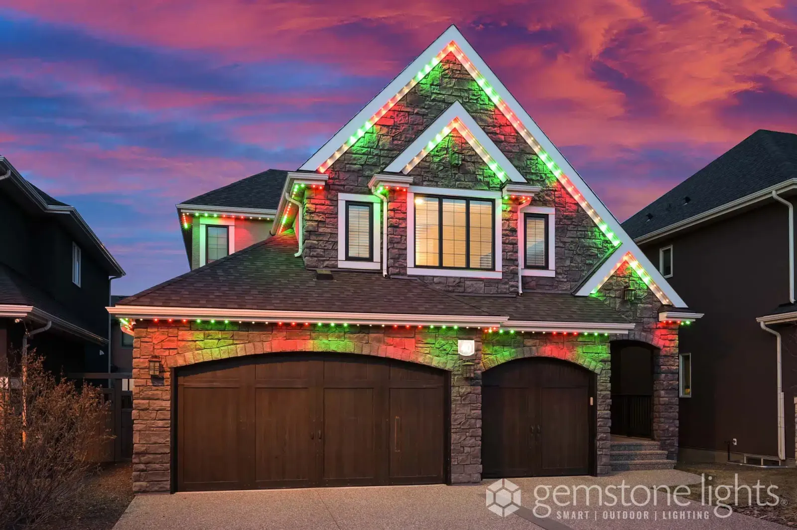 Stone home with tall gables lit in red, green and white Christmas lights under a pink evening sky