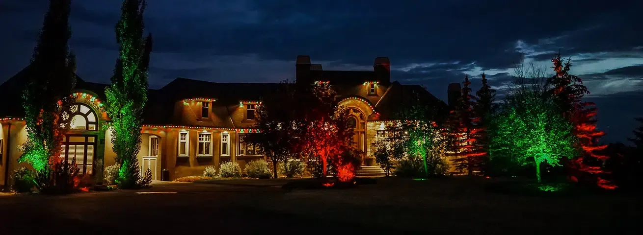 Stone house at night with roofline lights and tall trees uplit in red and green on either side