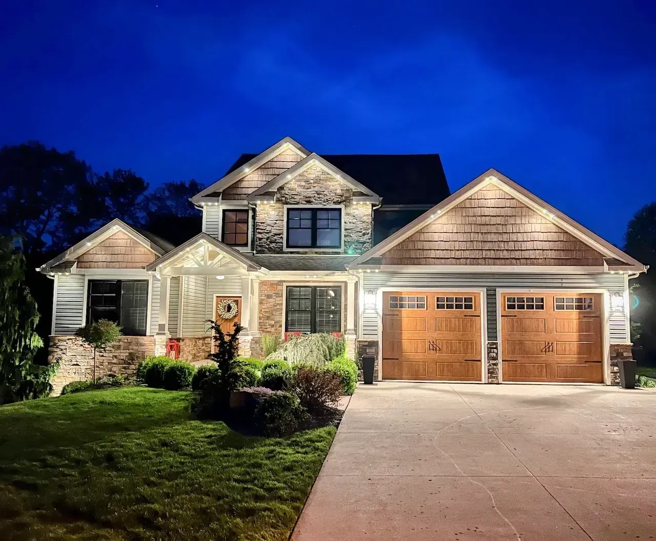 Two-story house at blue hour with bright roofline lights over a long driveway and front lawn